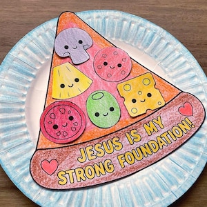 May include: A paper craft pizza slice featuring colorful cartoon toppings including a mushroom, pepperoni, pineapple, olive, and cheese. The crust area displays the text "JESUS IS MY STRONG FOUNDATION!" with two small pink hearts, all resting on a blue-rimmed paper plate.