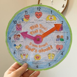 May include: A circular paper craft clock with a green border, blue colored background, and two movable orange and pink hands. The clock face features illustrations of hearts, hands, and feet with text including Tick Tock Tick Tock and I can trust God's clock.