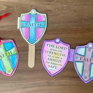 Op de afbeelding: Drie papieren schildvormige uitknipsels met kleurpotloodontwerpen. Eén schild heeft een rood lint, één is bevestigd aan een houten stokje en één is een vouwkaart met de tekst: THE LORD gives me STRENGTH. He is like a SHIELD that keeps me SAFE. PSALM 28:7.