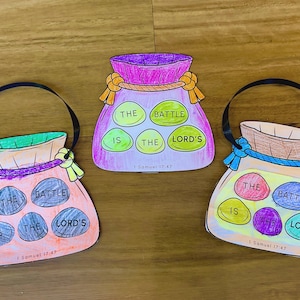 May include: Three paper cutouts shaped like pouches, each featuring five coloured stones with the text THE BATTLE IS THE LORD'S and 1 Samuel 17:47. The pouches are coloured in shades of pink, purple, and tan, with black ribbon handles attached.