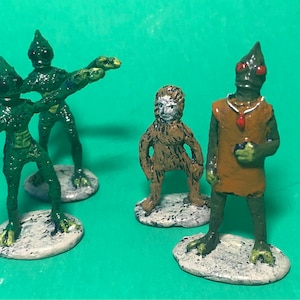 May include: Four painted figurines, two green aliens with guns, a brown ape-like creature, and a green alien with a red jewel necklace. The figurines are standing on white bases.