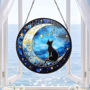 May include: A circular stained glass suncatcher featuring a black cat silhouette sitting on a textured yellow crescent moon. The background is a deep blue sky with scattered gold stars hanging from thin lines, all enclosed in a black frame.