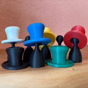 Könnte beinhalten: Eine Sammlung kleiner, zylinderförmiger Spielfiguren in verschiedenen Farben wie Weiß, Blau, Gelb, Rot, Grün und Schwarz. Jedes Teil hat eine breite Krempe und eine zylindrische Krone, steht aufrecht auf einer flachen Basis und ist gruppiert.