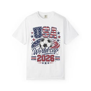 Pode incluir: Uma t-shirt branca de manga curta com um design gráfico contendo o texto USA World cup 2026. O design inclui uma bola de futebol, estrelas vermelhas e azuis e riscas vermelhas e azuis num estilo desgastado, centrado na frente da t-shirt.