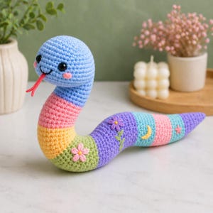 May include: A crocheted snake toy featuring a multi-colored striped body in blue, pink, yellow, green, and purple. The snake has black safety eyes, pink cheeks, and a red yarn tongue. Embroidered details include a flower, a moon, and a star.