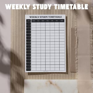 May include: A white weekly study timetable with black grid lines and the text "WEEKLY STUDY TIMETABLE". The timetable is on a textured beige surface with a shadow.