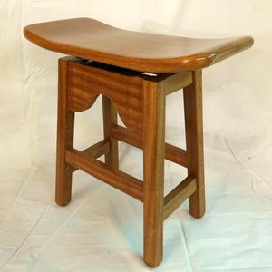 May include: A wooden stool with a curved, saddle-shaped seat and a simple, sturdy base. The stool is made of light brown wood and has a natural finish.