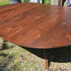 May include: A large, round dining table crafted from dark brown wood with a visible natural grain pattern. The tabletop features a smooth, polished surface and is supported by angled wooden legs extending from the center underneath the table.