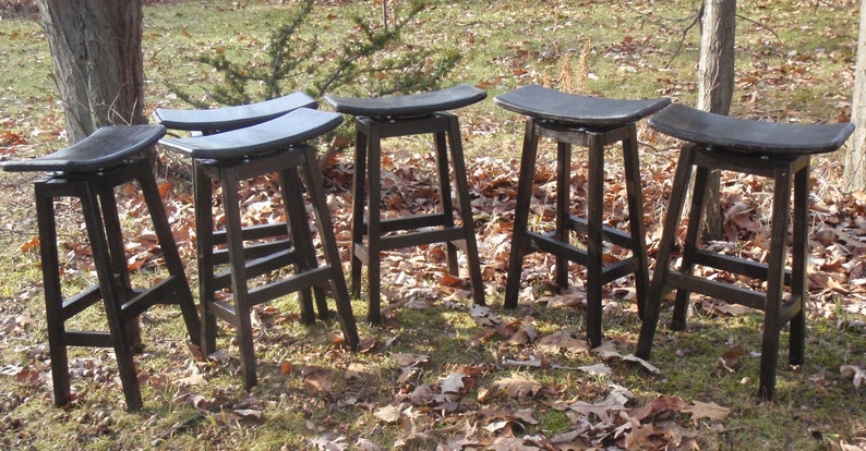 K&ouml;nnte beinhalten: Sechs schwarze Barhocker aus Holz mit geschwungenen Sitzen. Die Hocker sind in einer Reihe auf einer grasbewachsenen Fl&auml;che mit heruntergefallenen Bl&auml;ttern angeordnet.