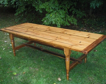 May include: A long, rectangular wooden table with a natural wood finish. The table has a simple design with turned legs and a stretcher. The table is shown outdoors on a grassy lawn.
