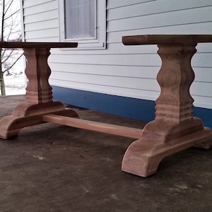 May include: A wooden table base with two pedestal legs and a connecting stretcher. The base is made of unfinished wood and has a natural color.