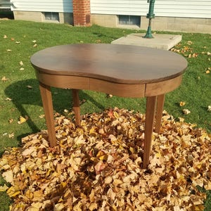 May include: A dark brown wooden desk with a curved top and a single drawer. The desk is sitting on a bed of fallen autumn leaves.
