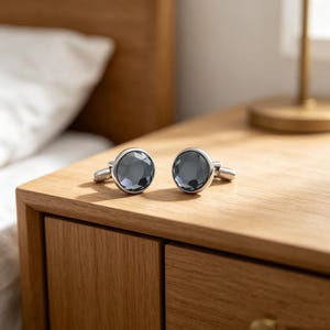 May include: A pair of round cufflinks featuring faceted dark gray stones set in polished silver-toned metal frames. Each cufflink has a circular face with a multi-faceted surface that reflects light, attached to a standard hinged toggle mechanism.