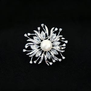 May include: A silver-toned brooch featuring a central white pearl surrounded by a starburst arrangement of marquise-cut clear stones and thin metal stems tipped with small, sparkling white crystals, creating a floral-inspired design.