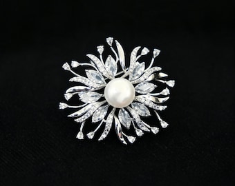 Può includere: Una spilla color argento con una perla bianca centrale circondata da una disposizione a raggiera di pietre trasparenti taglio marquise e sottili steli metallici con piccole punte di cristalli bianchi scintillanti.
