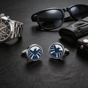 Puede incluir: Un par de gemelos plateados con un emblema azul y blanco, junto con un reloj de pulsera plateado, gafas de sol negras y una cartera de cuero negra. Los gemelos presentan un diseño circular con un pájaro estilizado.