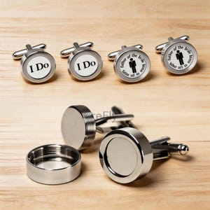 May include: Silver cufflinks with various inscriptions. Two pairs say "I Do" in black text on a white background. Two other pairs say "father of the bride" with a stick figure and text. The cufflinks are on a wooden surface.