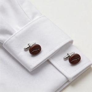 May include: Close-up of a white dress shirt with coffee bean-shaped cufflinks. The cufflinks are brown with silver-colored posts. The shirt's cuffs are neatly folded and buttoned.