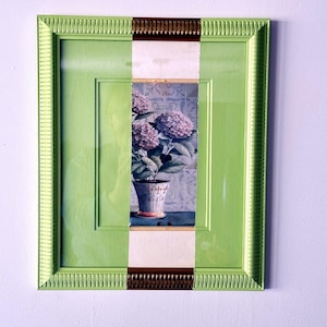Op de afbeelding: Een rechthoekig wandkunstwerk met een centraal verticaal paneel met een schilderij van paarse hortensia's in een decoratieve pot. De lijst is lichtgroen met twee horizontale bronskleurige accenten aan de boven- en onderkant.