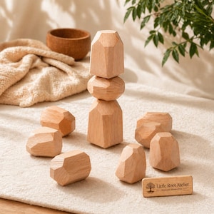 Könnte beinhalten: Ein Holz-Balance-Spielzeug-Set mit geometrischen Formen, darunter ein gestapelter Turm und mehrere einzelne Blöcke. Die Naturholzstücke sind hellbraun und liegen auf einer cremefarbenen Oberfläche. Ein kleines Holzschild trägt die Aufschrift "Little Root Atelier Montessori Wooden Toys."