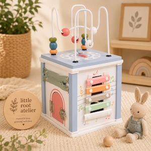 May include: A multi-activity wooden toy cube with a bead maze, xylophone, door, and zipper. The cube is painted in pastel colors and features the text "little root atelier". A small stuffed bunny sits nearby.