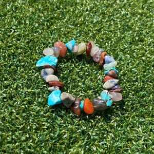 May include: A colorful bracelet made of various polished gemstones, including turquoise, orange, and brown stones. The bracelet is set against a backdrop of green grass, showcasing its natural beauty and vibrant colors. The bracelet is a unique piece of jewelry.
