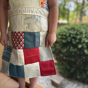 Puede incluir: Un delantal de patchwork con un babero y una cinturilla texturizados de color crema. La falda del delantal presenta una variedad de cuadrados de tela en rojo, azul y crema, con diseños estampados y a rayas. El delantal lo lleva una persona.