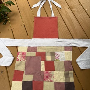 May include: A patchwork apron with a red top and a patterned bottom. The apron has a white neck strap and waist ties. The bottom of the apron is a patchwork of various fabrics in shades of red, pink, and beige.