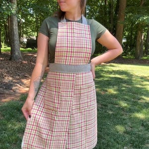 Op de afbeelding: Een vintage schort met een ruitpatroon in rood, groen en crème. De schort heeft een grijze tailleband en bandjes. De persoon op de foto draagt een groen t-shirt en witte sandalen.