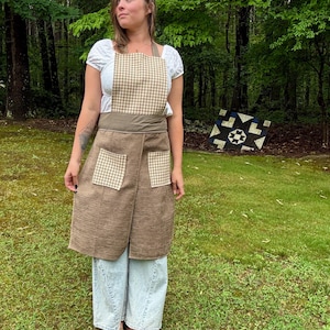 May include: A brown apron with a houndstooth pattern on the bib and pockets. The apron has a wrap-around design and is worn over a white top and light blue jeans. The background features a green lawn and trees.