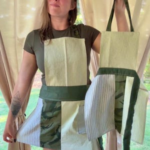 Könnte beinhalten: Eine Schürze und eine passende Tragetasche. Die Schürze hat ein Patchwork-Design mit grün-weiß gestreiftem Stoff, Camouflage-Print und einem dunkelgrünen Bund. Die Tragetasche hat den gleichen gestreiften Stoff und dunkelgrüne Träger. Die Schürze wird von einer Person hochgehalten.