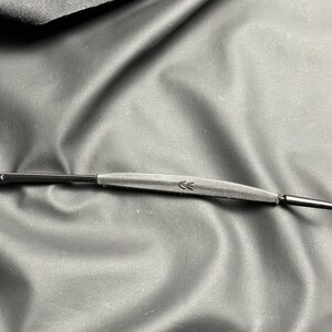May include: Close-up of a black eyeglass frame with a gray bridge and thin arms. The bridge features two small arrow symbols. The frame is set against a textured, dark background.