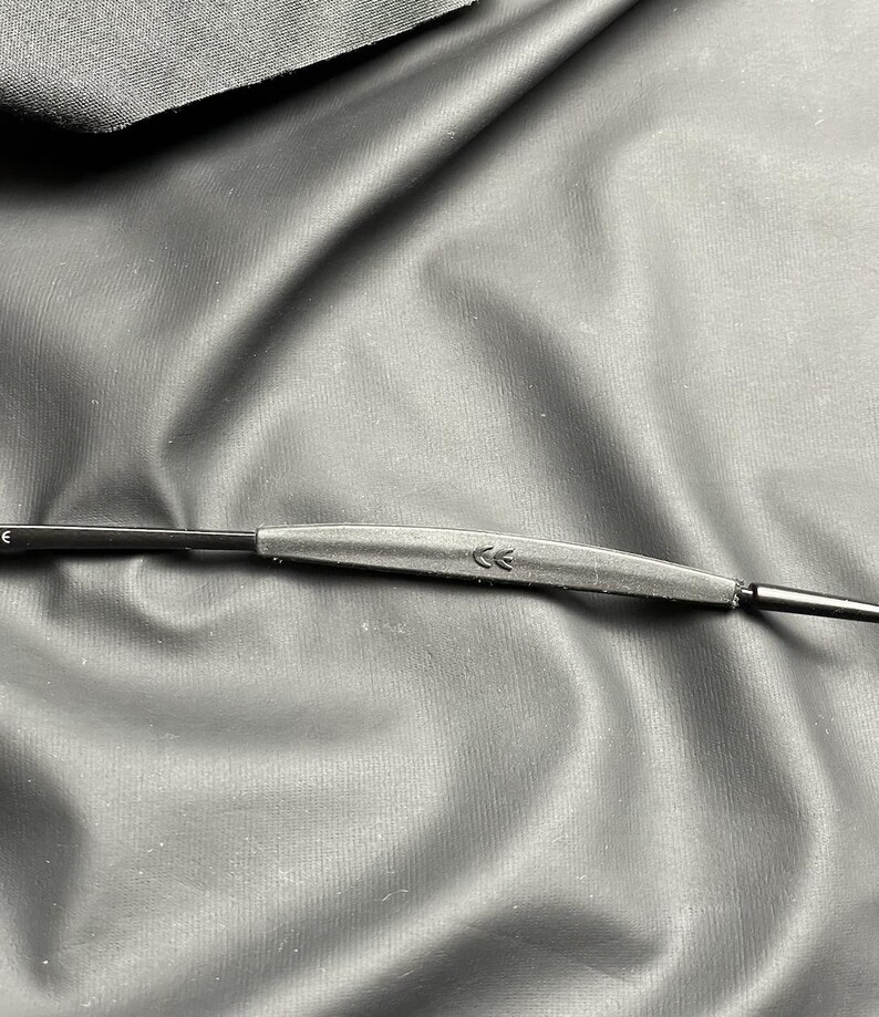 May include: Close-up of a black eyeglass frame with a gray bridge and thin arms. The bridge features two small arrow symbols. The frame is set against a textured, dark background.