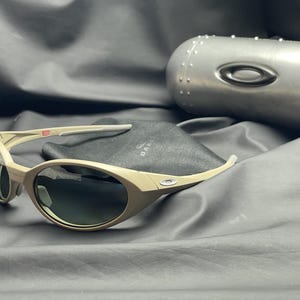 Puede incluir: Gafas de sol color beige con lentes oscuras y estuche a juego. Las gafas tienen un dise&ntilde;o curvo. El estuche es met&aacute;lico y de forma ovalada. Un peque&ntilde;o detalle de bandera es visible en la patilla.