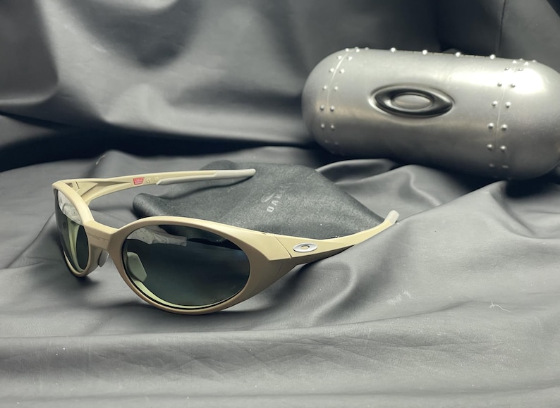 Puede incluir: Gafas de sol color beige con lentes oscuras y estuche a juego. Las gafas tienen un dise&ntilde;o curvo. El estuche es met&aacute;lico y de forma ovalada. Un peque&ntilde;o detalle de bandera es visible en la patilla.