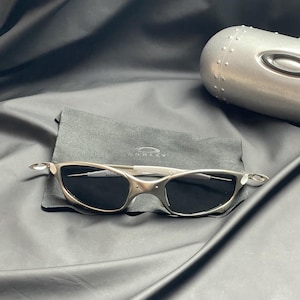 May include: Silver-toned sunglasses with dark lenses rest on a black cloth, accompanied by a matching case. The sunglasses have a sleek, modern design with a curved frame and a subtle logo. The case is metallic and has a riveted design.