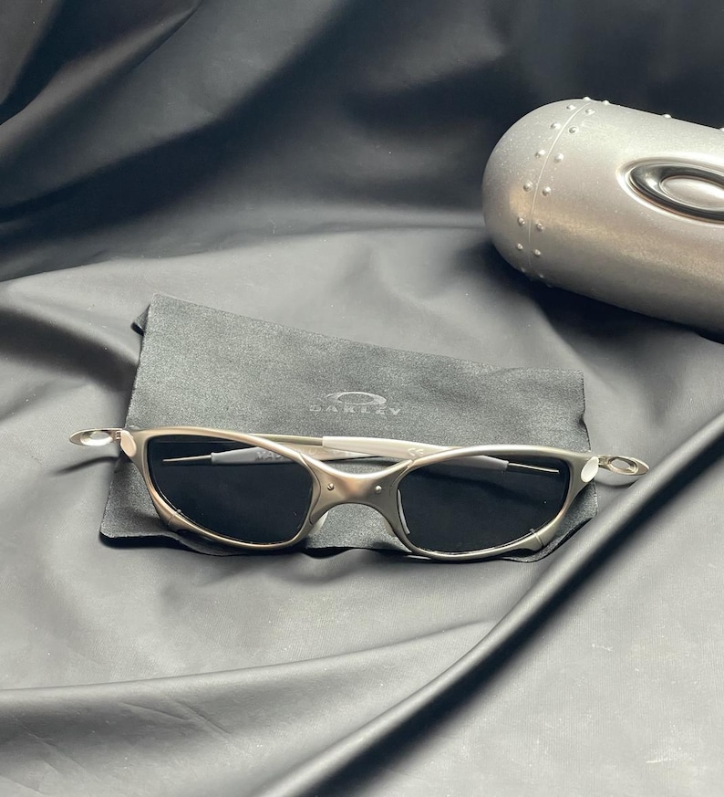 May include: Silver-toned sunglasses with dark lenses rest on a black cloth, accompanied by a matching case. The sunglasses have a sleek, modern design with a curved frame and a subtle logo. The case is metallic and has a riveted design.