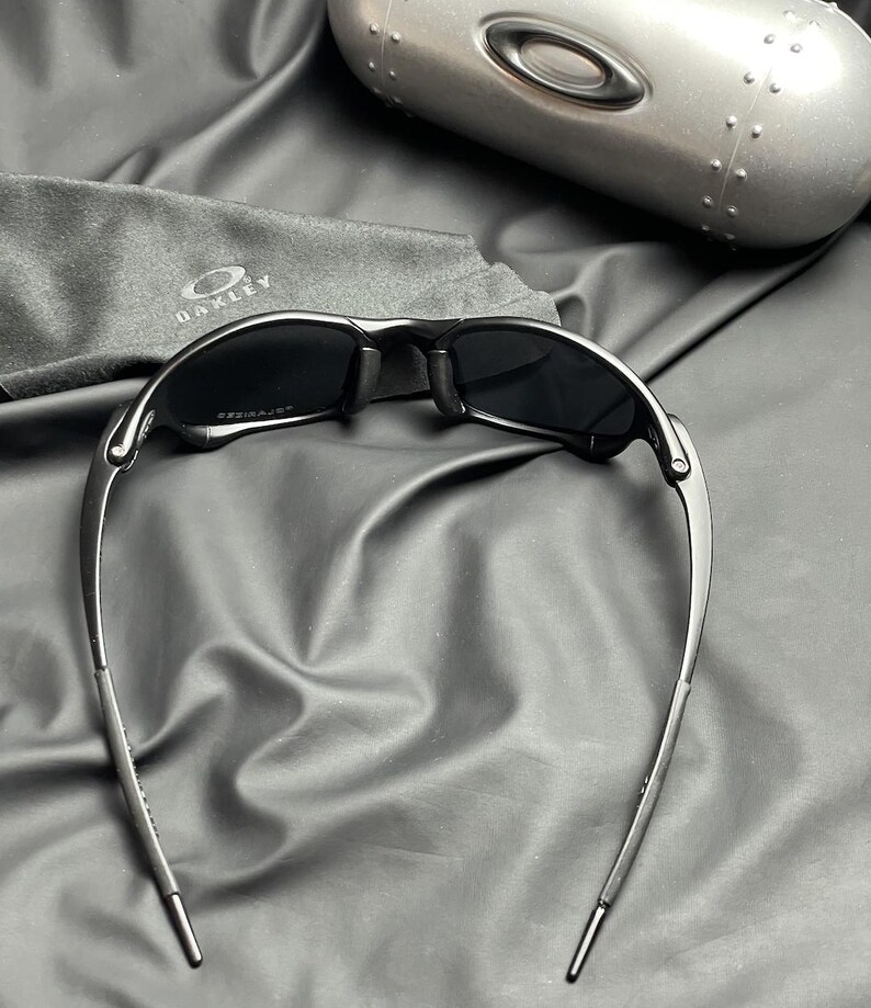 May include: Black sunglasses with a sleek, modern design. The sunglasses have a dark frame and lenses, with a curved shape. A gray case and cleaning cloth with the Oakley logo are also visible.