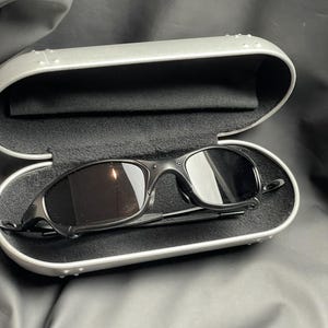 May include: Black sunglasses with a curved frame rest inside a silver case with a black interior. The sunglasses have dark lenses and a sleek, modern design. The case is open, revealing the sunglasses.