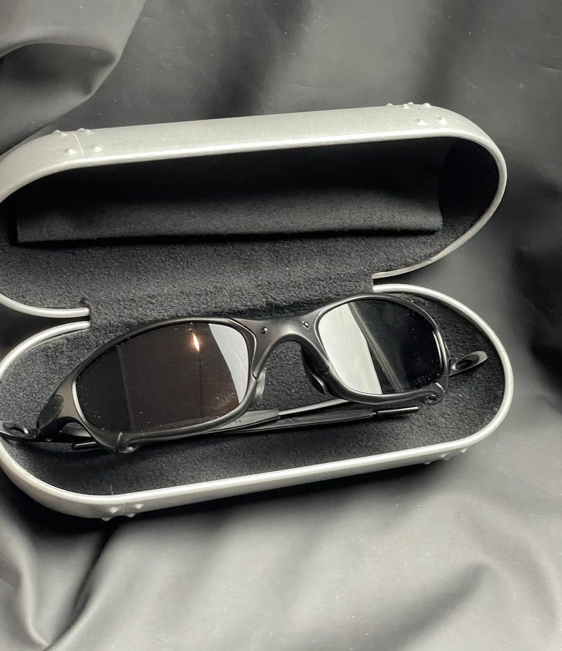 May include: Black sunglasses with a curved frame rest inside a silver case with a black interior. The sunglasses have dark lenses and a sleek, modern design. The case is open, revealing the sunglasses.