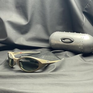 Puede incluir: Gafas de sol Oakley beige con lentes oscuras, sobre una tela negra. Las gafas tienen una montura curvada y van acompa&ntilde;adas de un estuche plateado con el logotipo de Oakley.