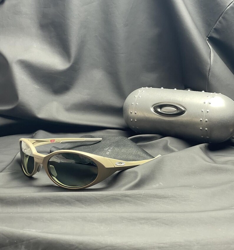 Puede incluir: Gafas de sol Oakley beige con lentes oscuras, sobre una tela negra. Las gafas tienen una montura curvada y van acompa&ntilde;adas de un estuche plateado con el logotipo de Oakley.