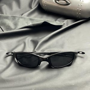 May include: Black sunglasses with a sleek, curved design, featuring dark lenses and a matching black frame. The image also shows a silver case and a black cleaning cloth, all set against a dark background.