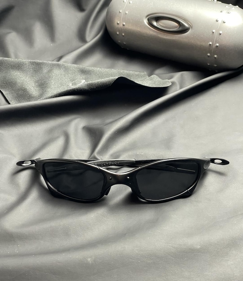 May include: Black sunglasses with a sleek, curved design, featuring dark lenses and a matching black frame. The image also shows a silver case and a black cleaning cloth, all set against a dark background.