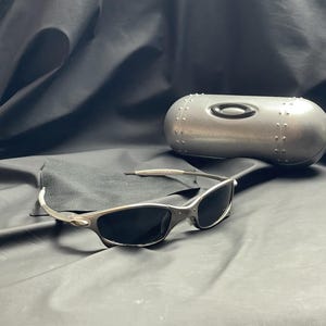 May include: Silver-framed sunglasses with dark lenses rest on a black cloth, accompanied by a matching silver case. The sunglasses have a sleek, modern design, and the case features a rivet-like pattern.