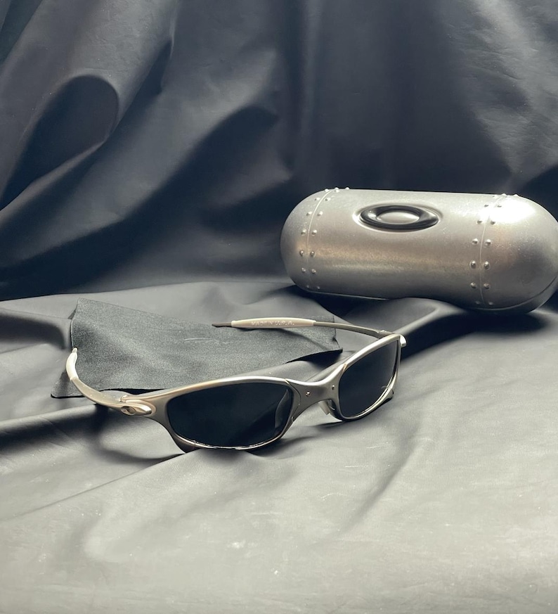 May include: Silver-framed sunglasses with dark lenses rest on a black cloth, accompanied by a matching silver case. The sunglasses have a sleek, modern design, and the case features a rivet-like pattern.