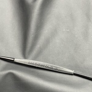 May include: A black tool with the text "MADE IN U.S.A." embossed on a rubberized grip. The tool has a thin, pointed end on either side. The background is a dark, textured surface.
