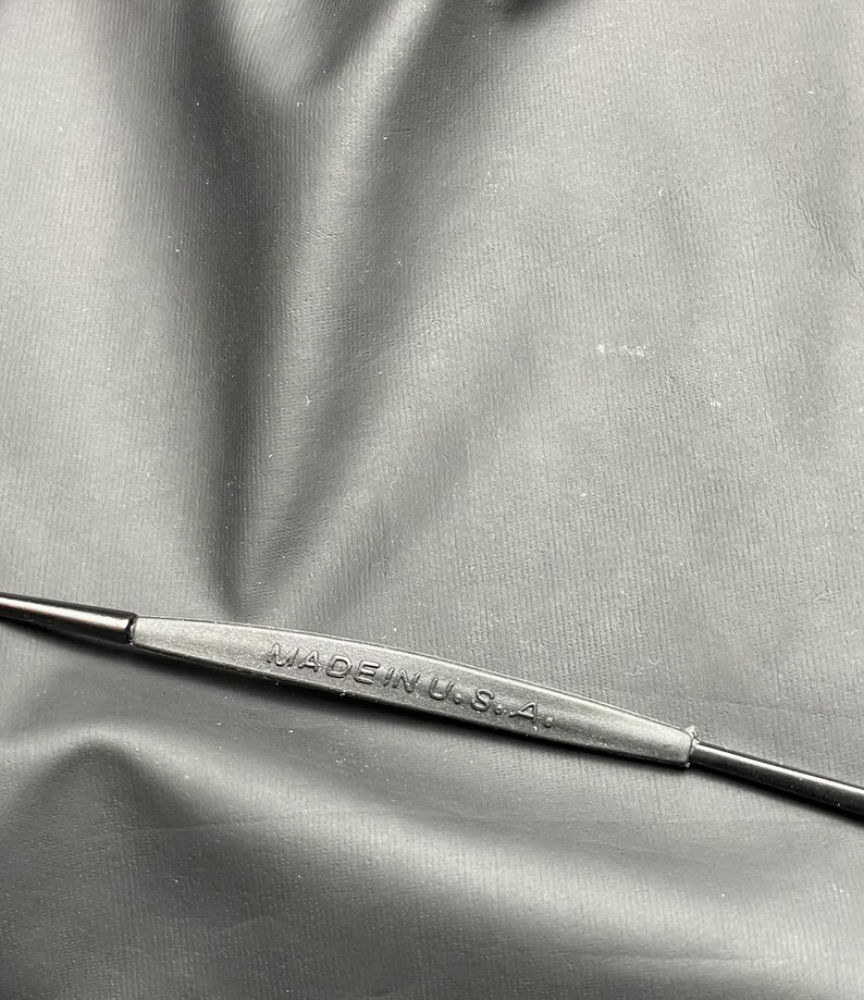May include: A black tool with the text "MADE IN U.S.A." embossed on a rubberized grip. The tool has a thin, pointed end on either side. The background is a dark, textured surface.
