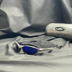 May include: Silver-framed sunglasses with blue lenses rest on a black cloth. A silver case with a black oval logo is in the background. The sunglasses have a sleek, modern design.