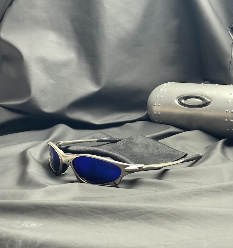 May include: Silver-framed sunglasses with blue lenses rest on a black cloth. A silver case with a black oval logo is in the background. The sunglasses have a sleek, modern design.